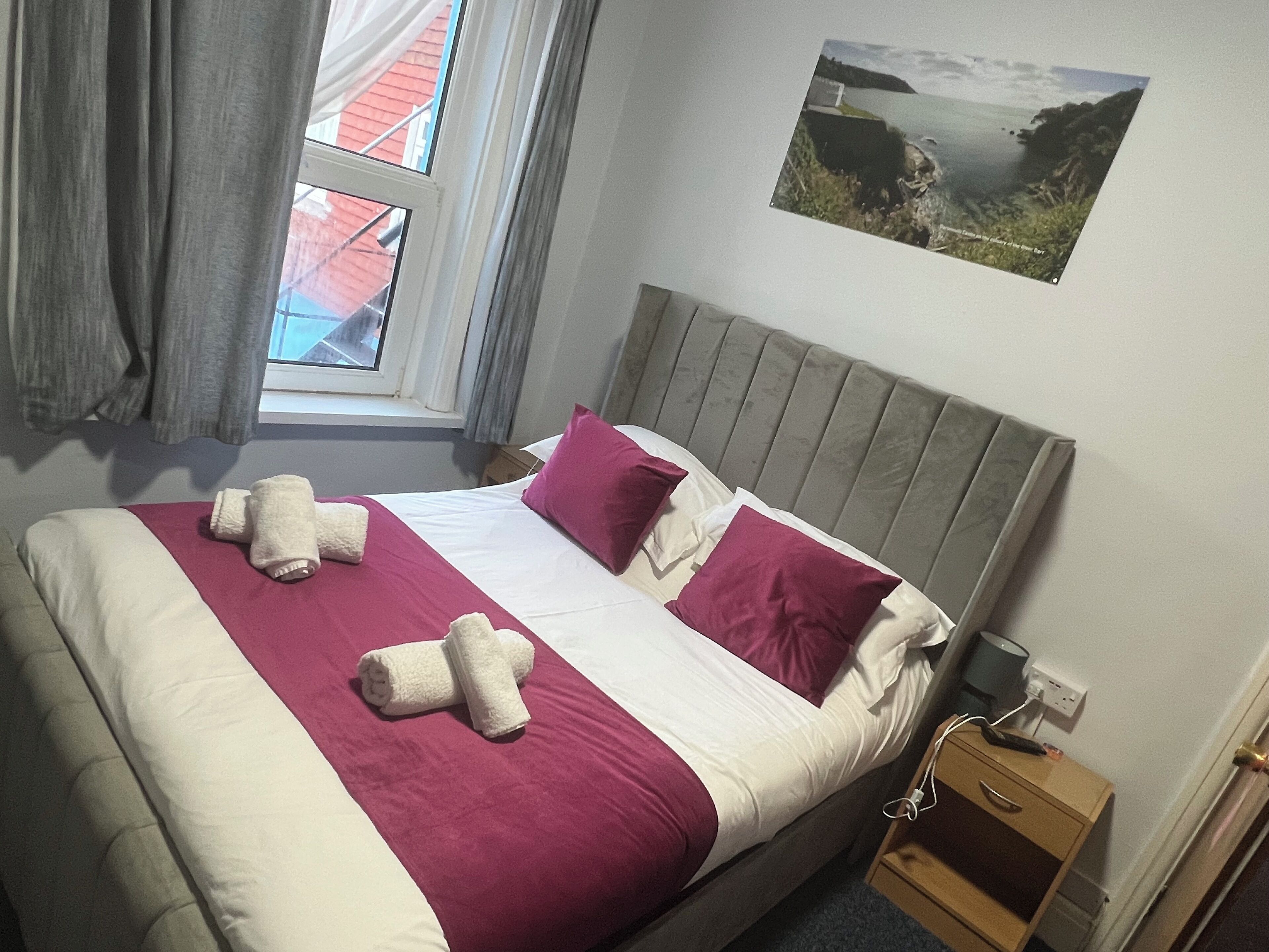 A bed with a headboard, pillows, and a blanket. There is a window with curtains, a picture on the wall, and a nightstand with a lamp.