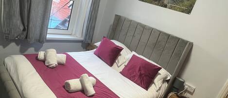 A bed with a headboard, pillows, and a blanket. There is a window with curtains, a picture on the wall, and a nightstand with a lamp.