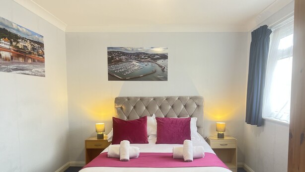 Cama bem arrumada com cabeceira estofada, duas luminárias de apoio e um quadro emoldurado acima.