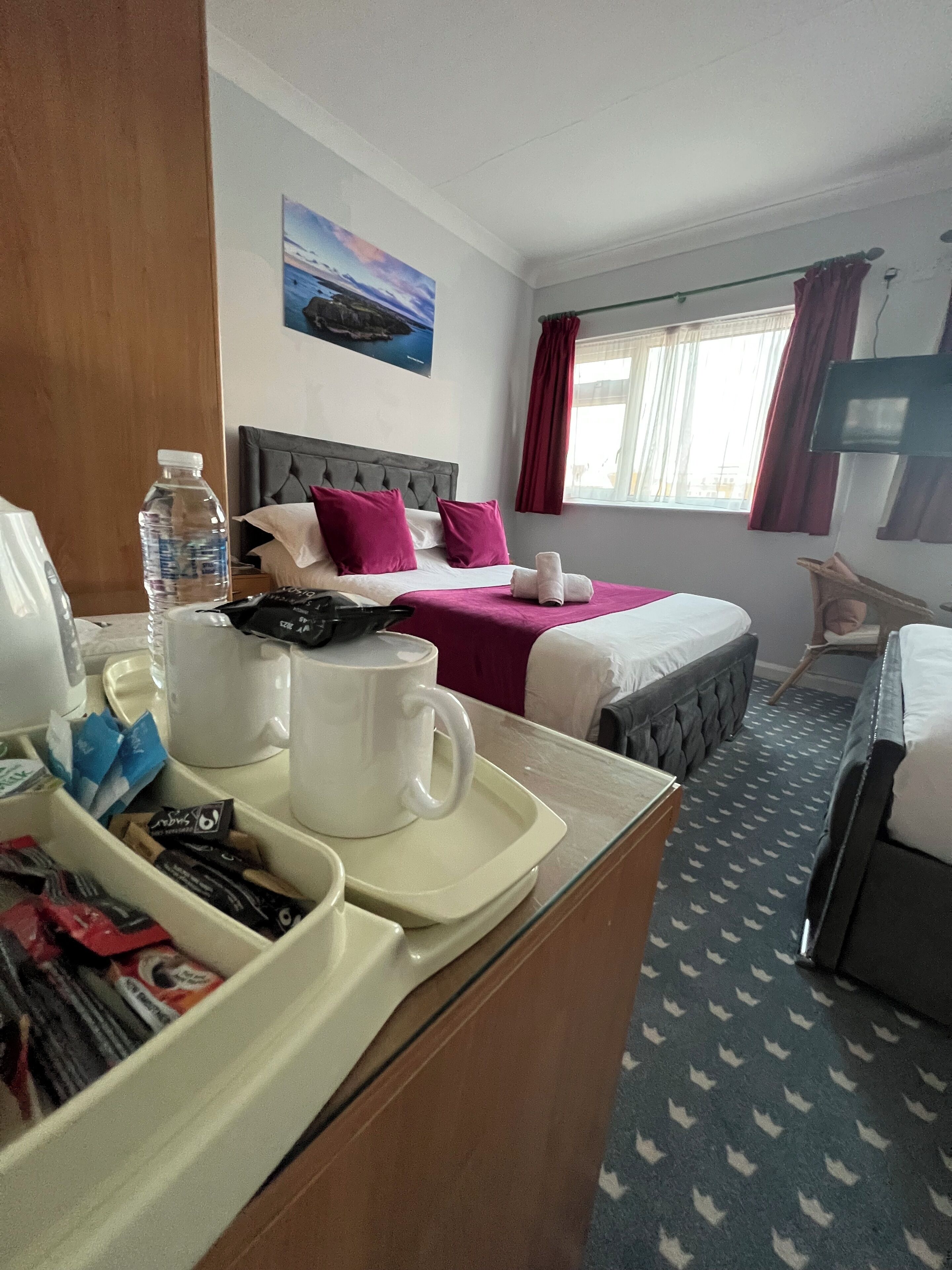 A hotel room with two beds, a desk with a tray of breakfast items, a window with curtains, and a painting on the wall.