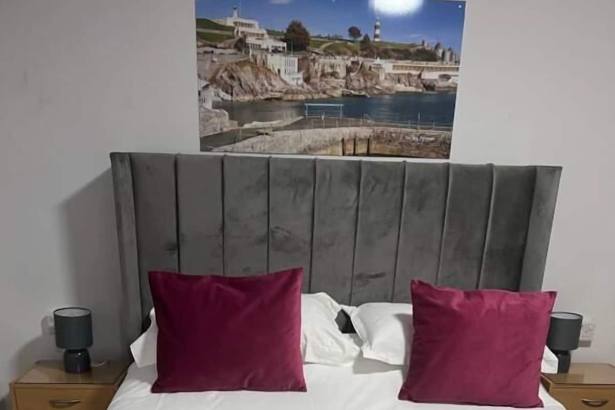 A neatly made bed with a white and maroon bedspread, two maroon pillows, and two white pillows. There are two nightstands with lamps, and a framed picture of a coastal scene hangs above the bed.