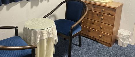 A room with a blue chair, a small round table with a lace tablecloth, a wooden dresser with drawers, and a framed picture of a coastal scene.
