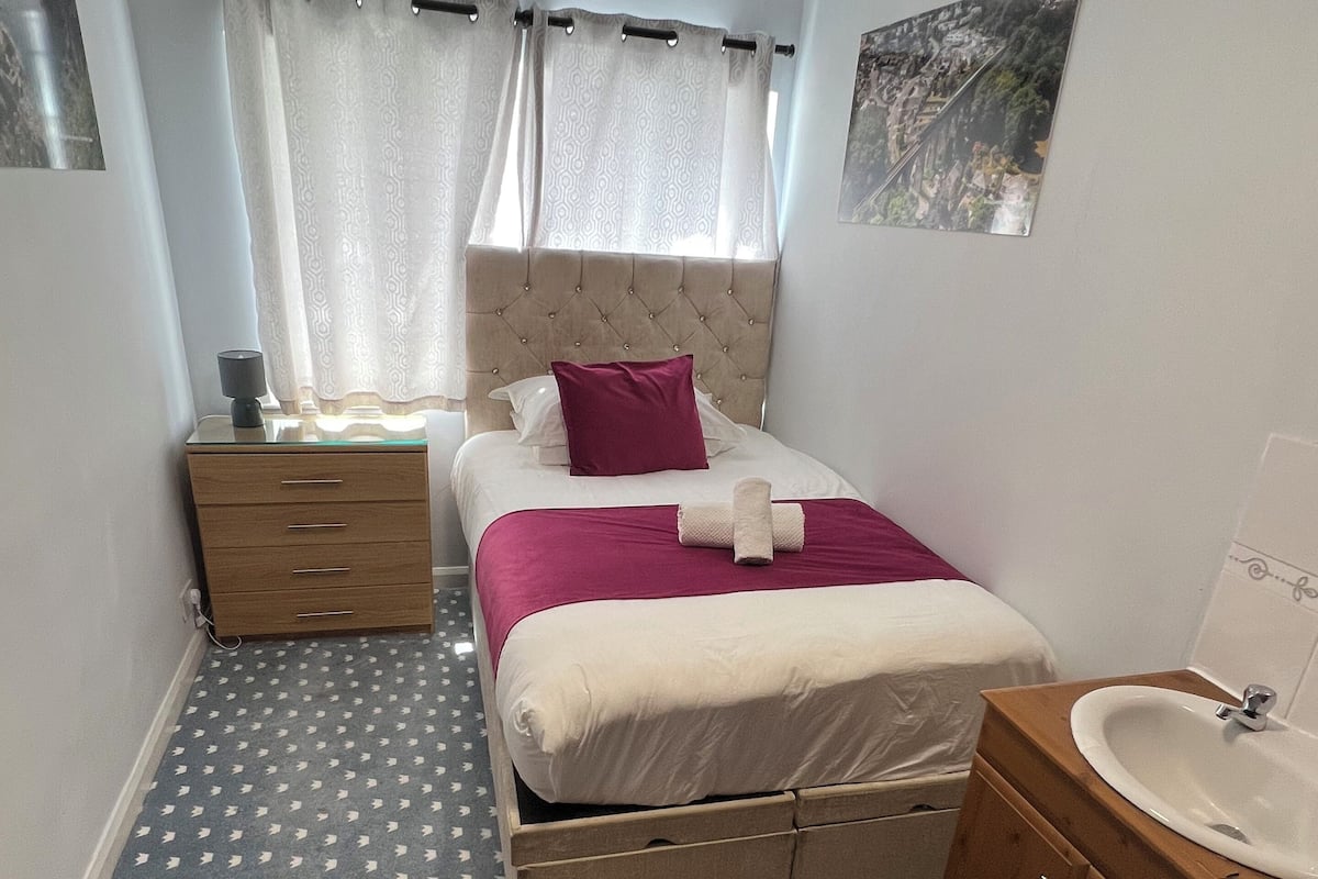 A bedroom with a bed, a nightstand, a dresser, a bathroom with a sink, and a window with curtains.
