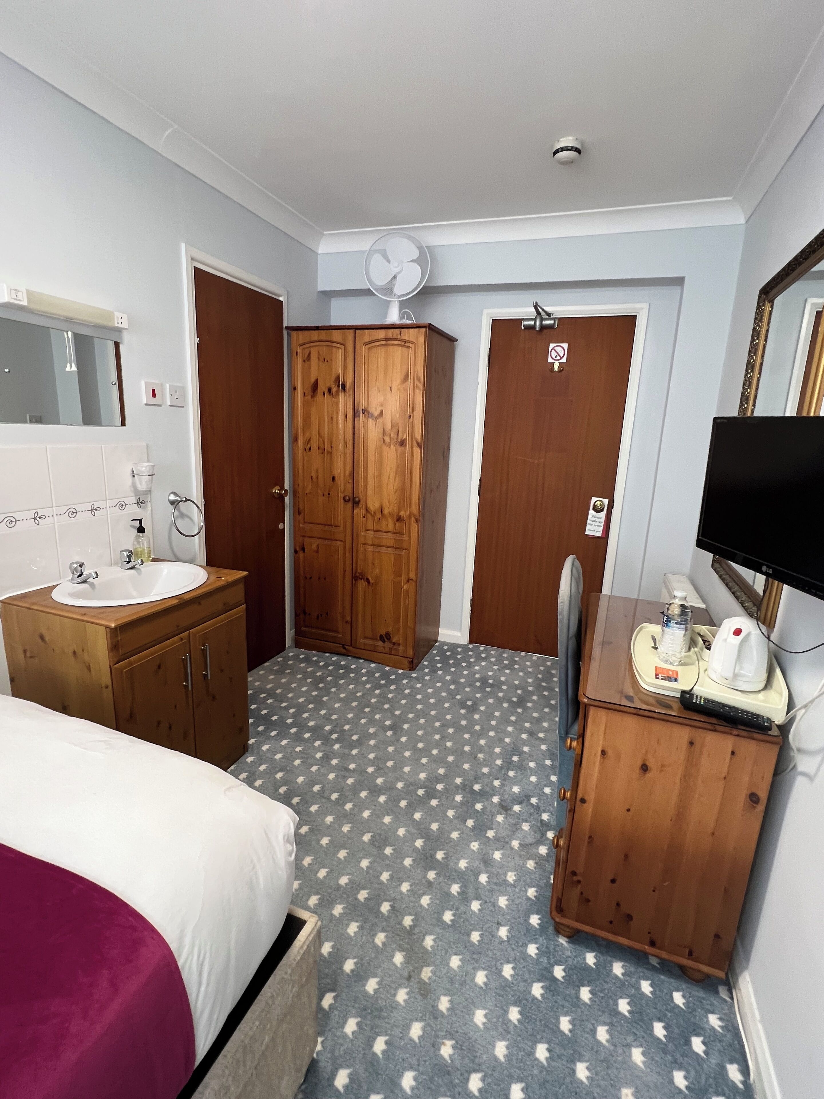 Habitación de hotel con cama, armario de madera, pequeño escritorio con televisor, mesita de noche con lámpara y baño con lavamanos y espejo.