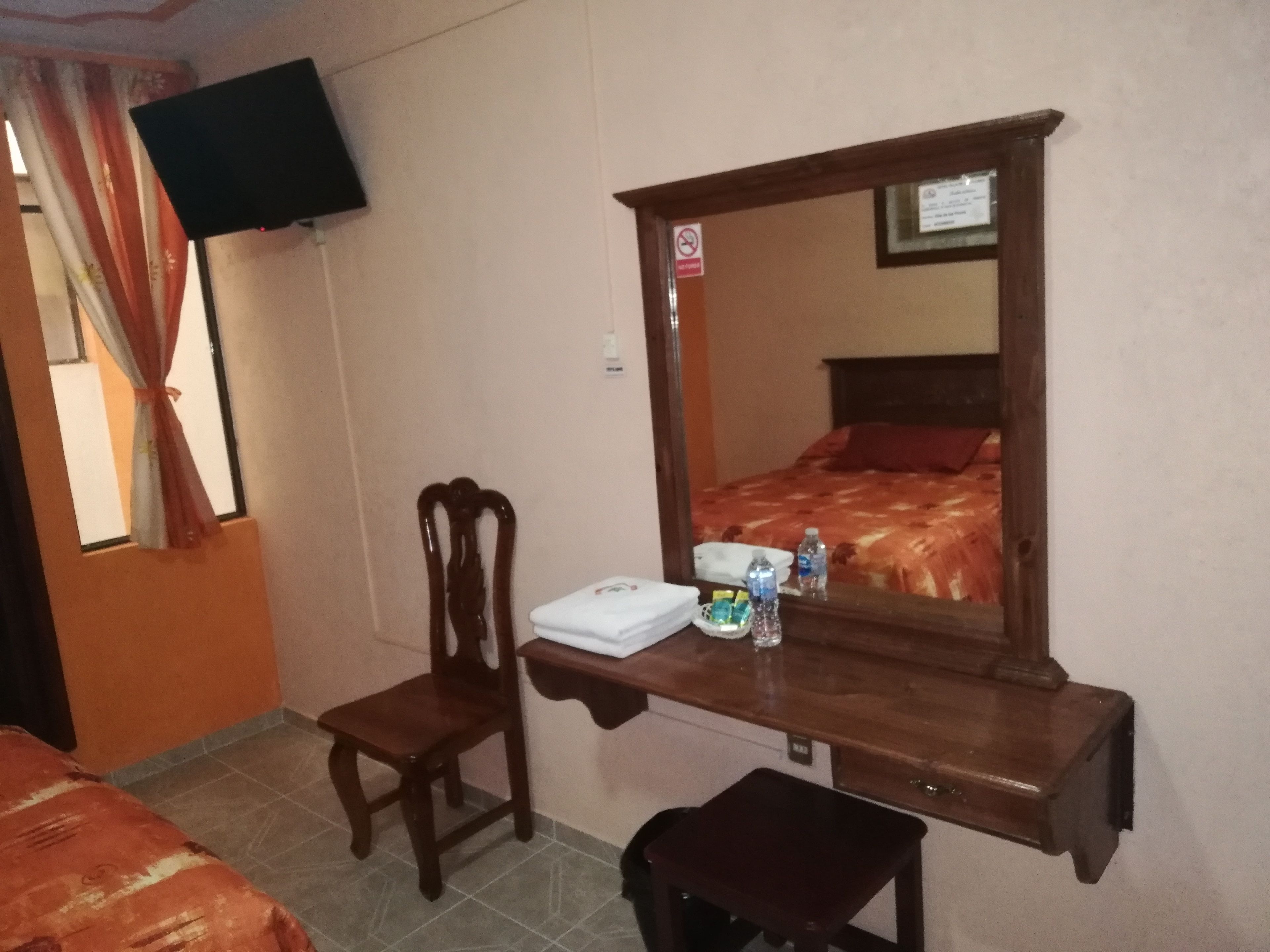 A hotel room with a bed, a wooden desk with a mirror, a chair, a television mounted on the wall, and a window with curtains.