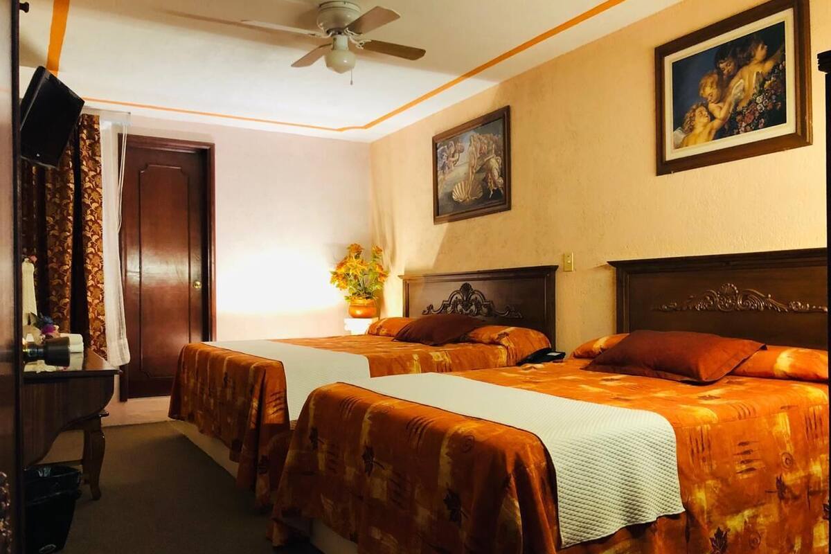 Habitación de hotel con dos camas, ventilador de techo, un cuadro y un escritorio con lámpara.