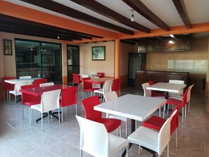 Um restaurante com mesas e cadeiras vermelhas e brancas, uma área de cozinha e janelas amplas.