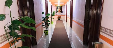Un couloir d’hôtel avec un tapis rouge, des plantes en pot et une poubelle.