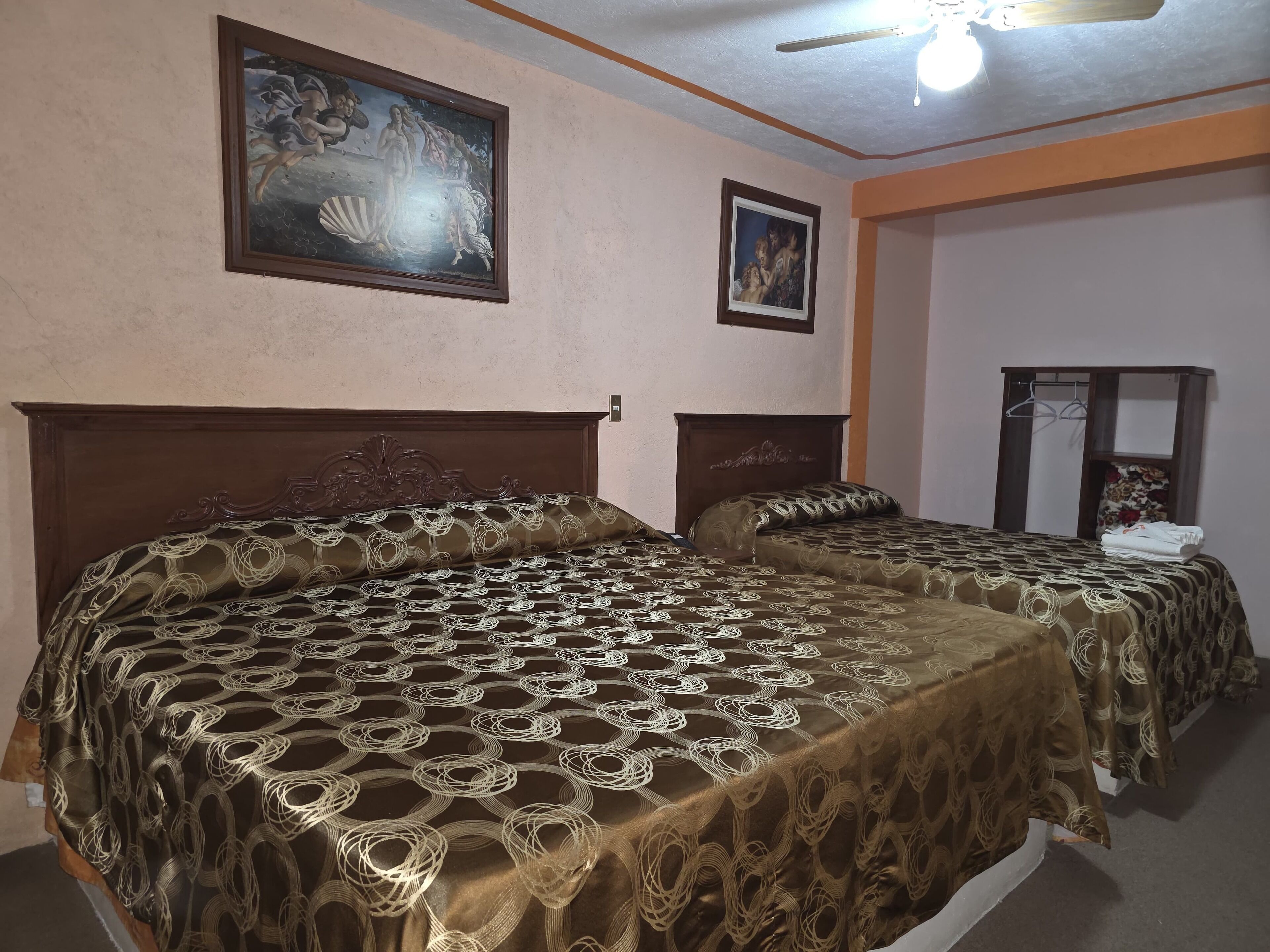 Habitación de hotel con dos camas, cada una con una colcha estampada, un ventilador de techo y dos cuadros enmarcados en las paredes.