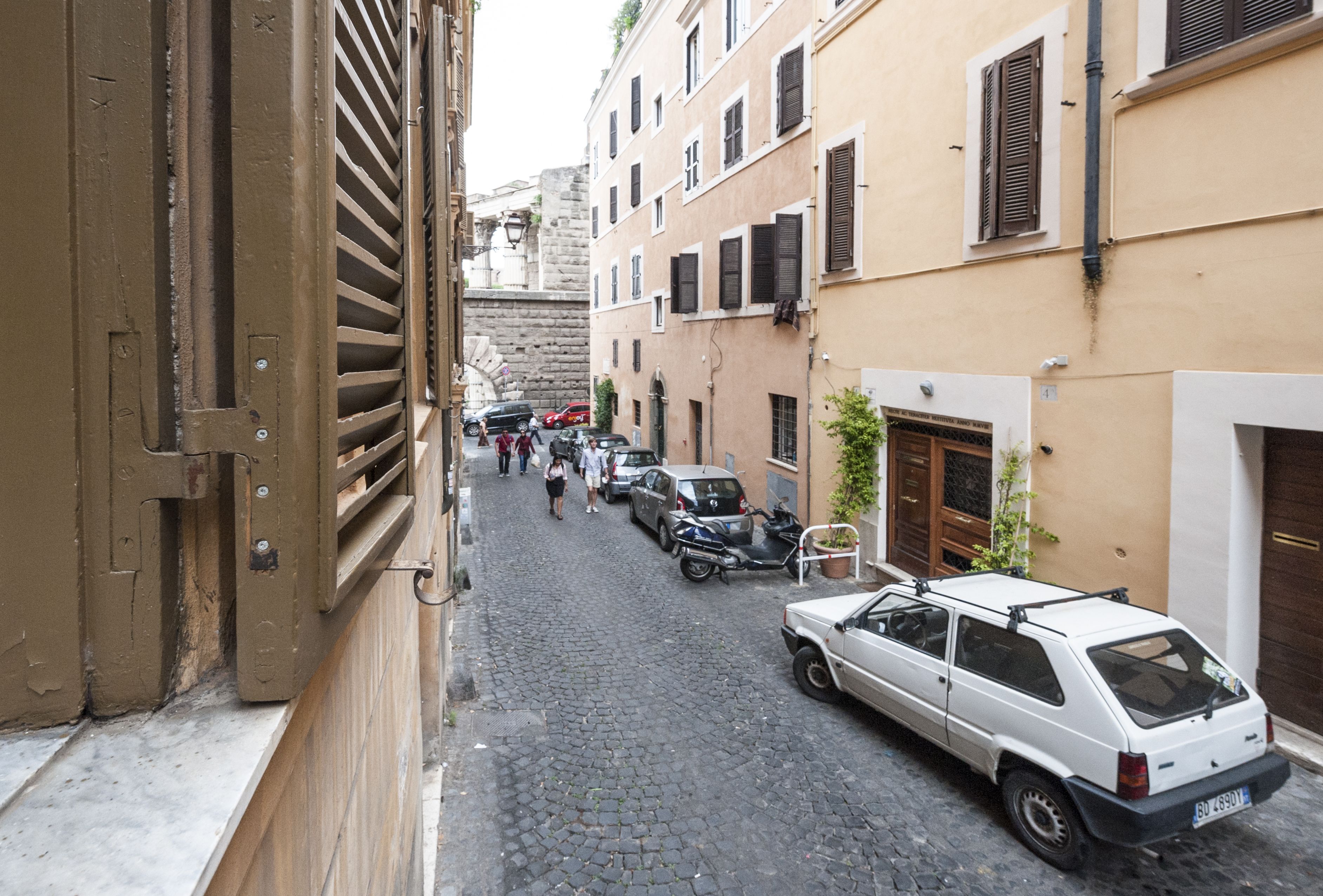 A narrow cobblestone street lined with buildings, parked cars, and a few pedestrians.