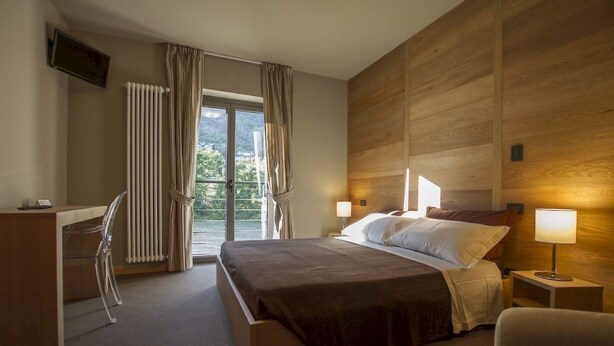 A hotel room with a bed, a desk, a chair, a TV, a window with curtains, and a wooden wall. - Chambres D'Hotes La Bicoque (Sarre)