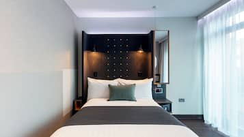 A modern hotel room with a large bed, a headboard with illuminated buttons, a bedside table with a lamp, and a large mirror.