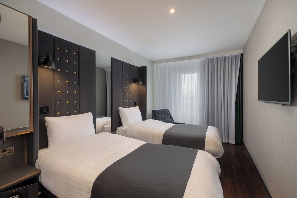 A hotel room with two beds, a TV, a desk, and a large window with curtains.