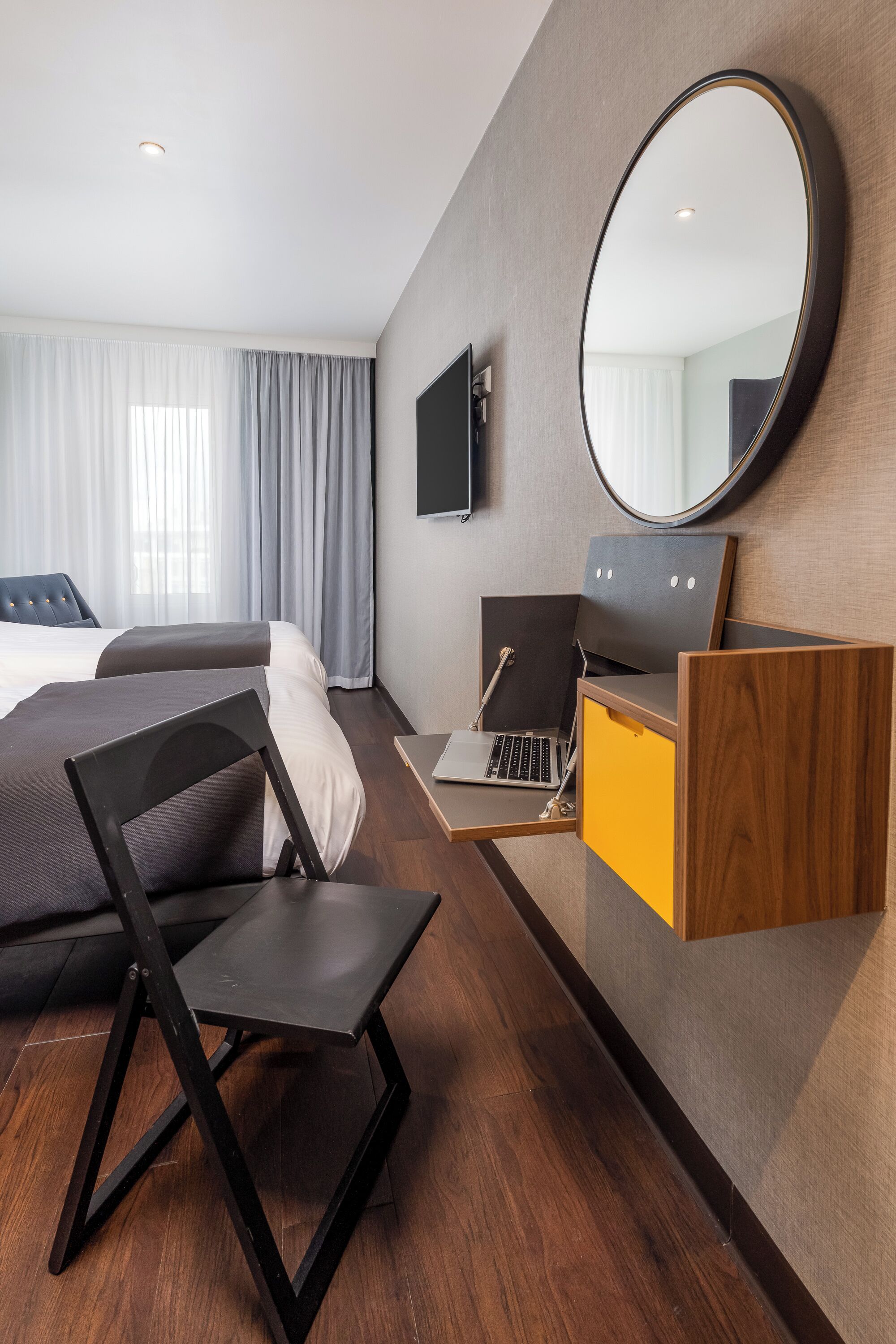 A hotel room with a bed, a desk with a laptop, a chair, a mirror, and a television.