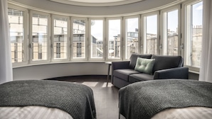Une chambre moderne avec une baie vitrée incurvée, un canapé gris, une petite table et deux lits recouverts de couvertures grises.