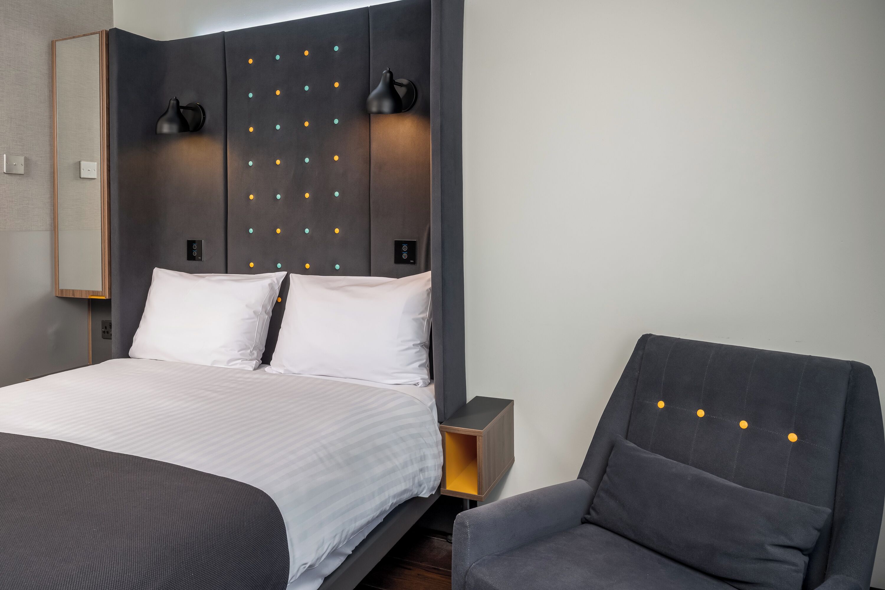 Una habitación de hotel moderna con una cama grande, una mesita de noche, una silla y un cabecero con botones decorativos.