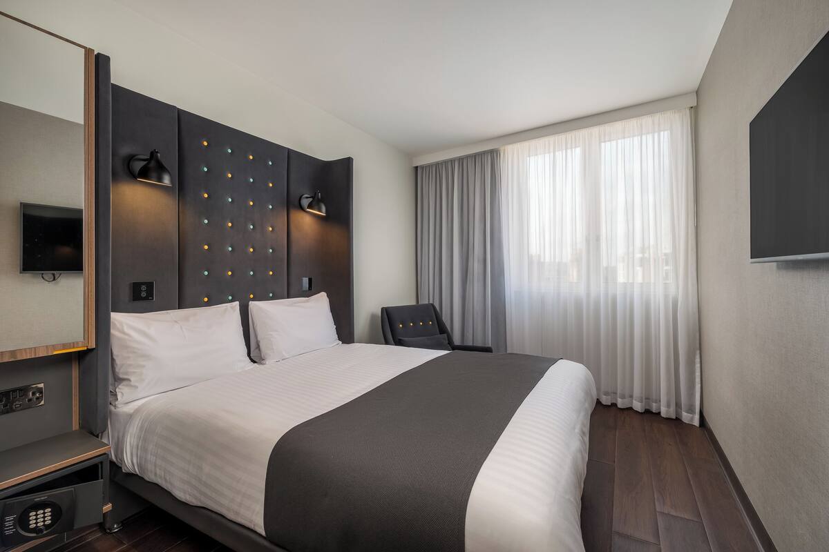 A modern hotel room with a large bed, a flat-screen TV, a desk with a lamp, and a chair.