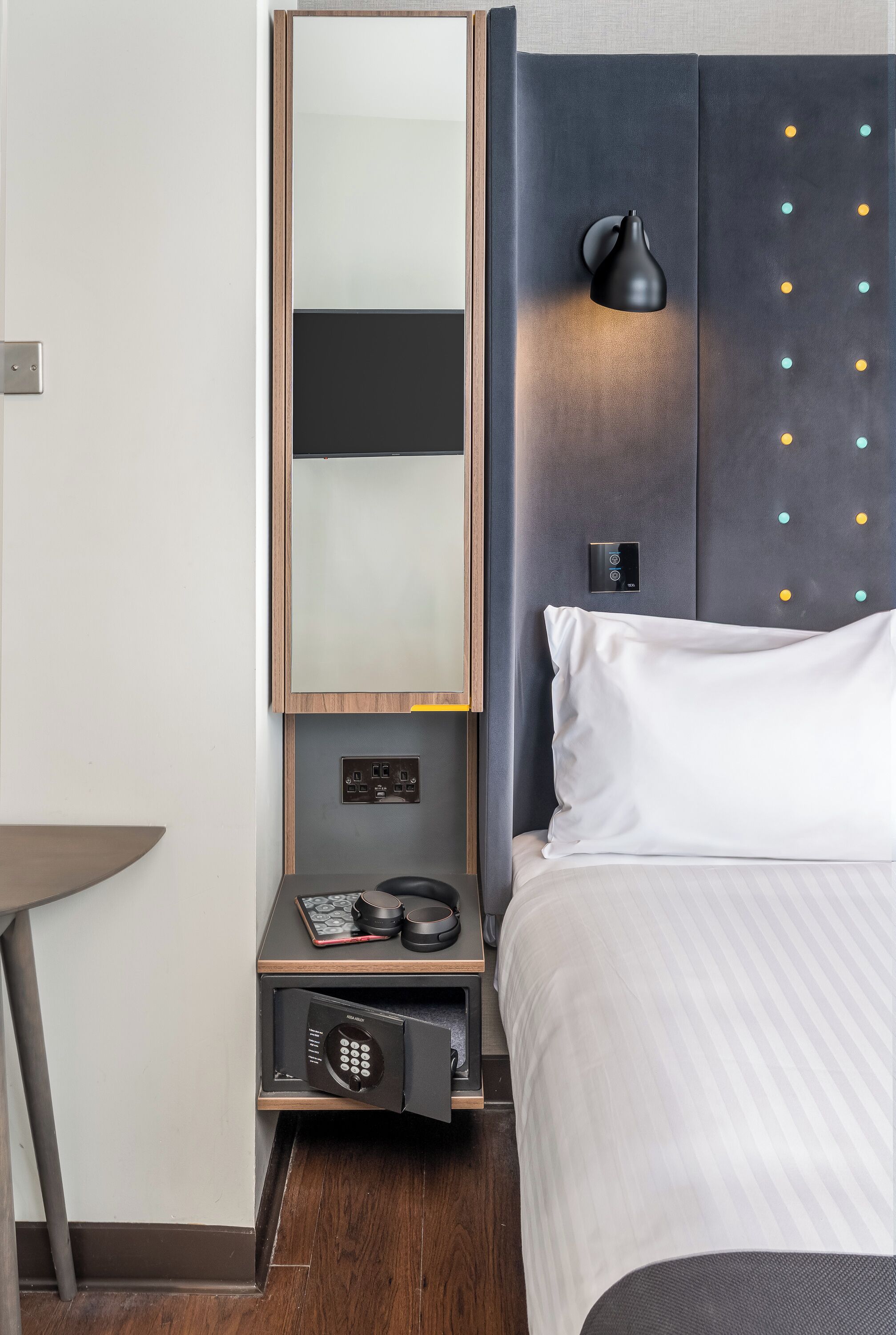 Une chambre d’hôtel équipée d’un lit, d’une table de chevet, d’un miroir, d’une lampe fixée au mur et d’un panneau de commande.