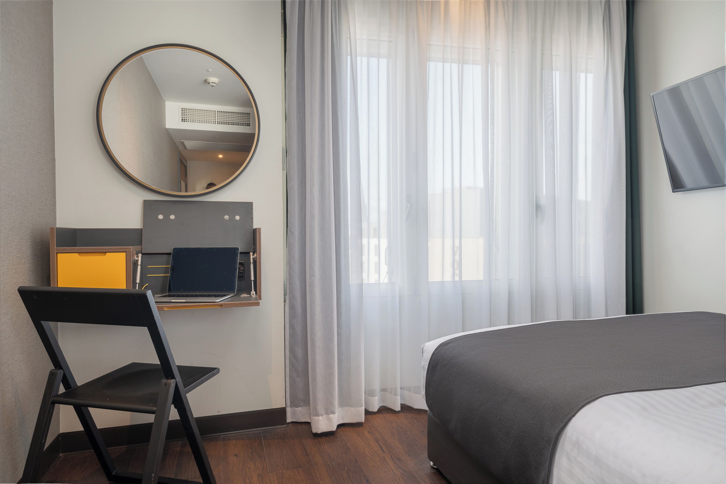 A hotel room with a bed, a desk with a laptop, a chair, a round mirror, and sheer curtains.