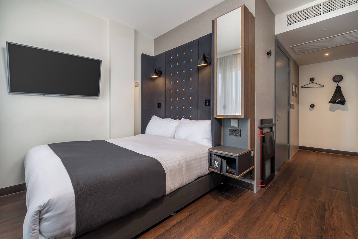 A modern hotel room with a large bed, a flat-screen TV, a bedside table, a mirror, and a wardrobe.