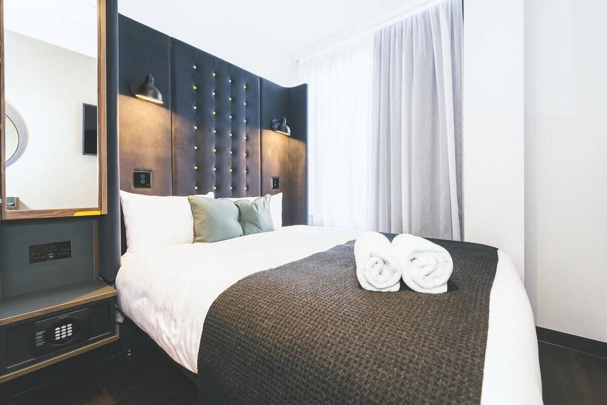 A hotel room with a large bed, a headboard with a light fixture, a bedside table, and a window with curtains.