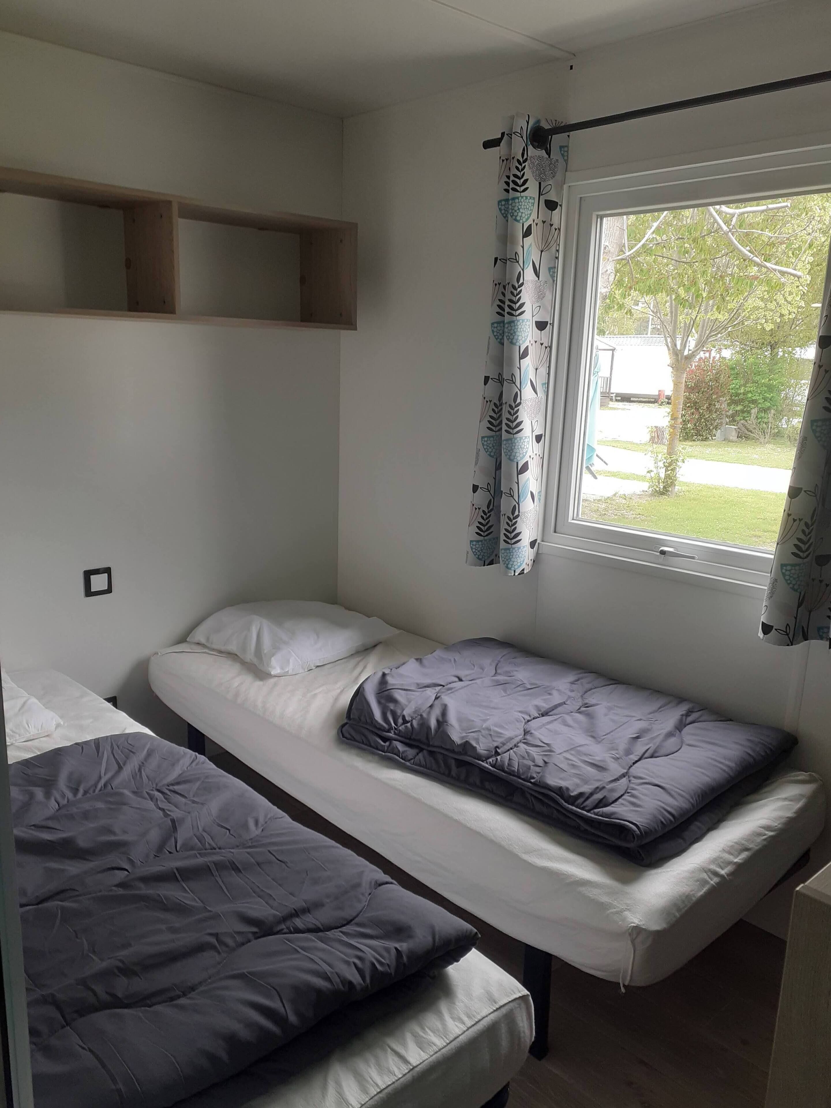 A dormitory room with two single beds, a window with curtains, and a wooden shelf above one bed.