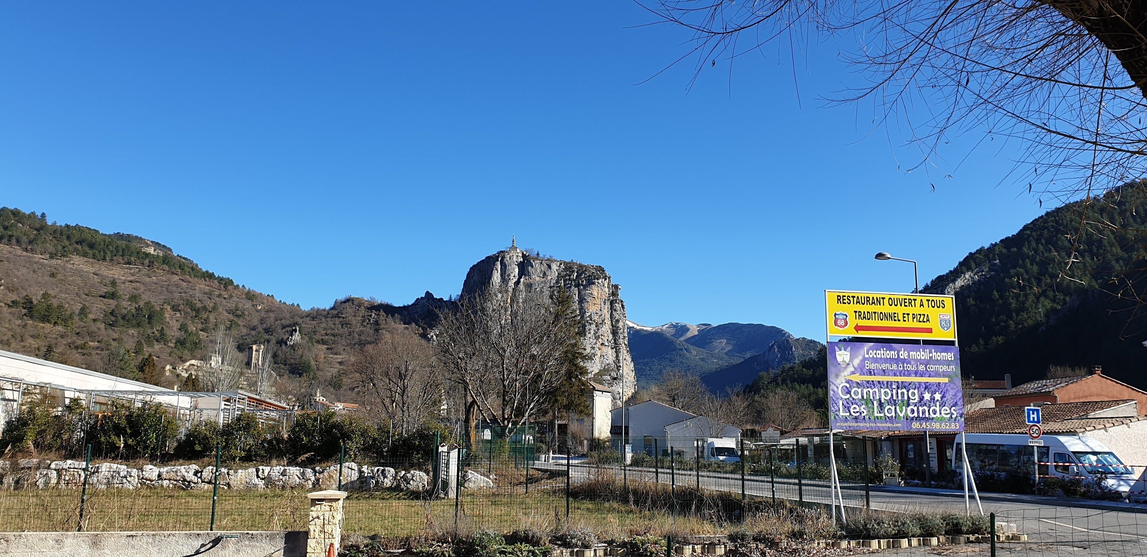 A mountainous landscape with a prominent rock formation, a road with vehicles, and a signboard advertising a restaurant and mobile home locations.