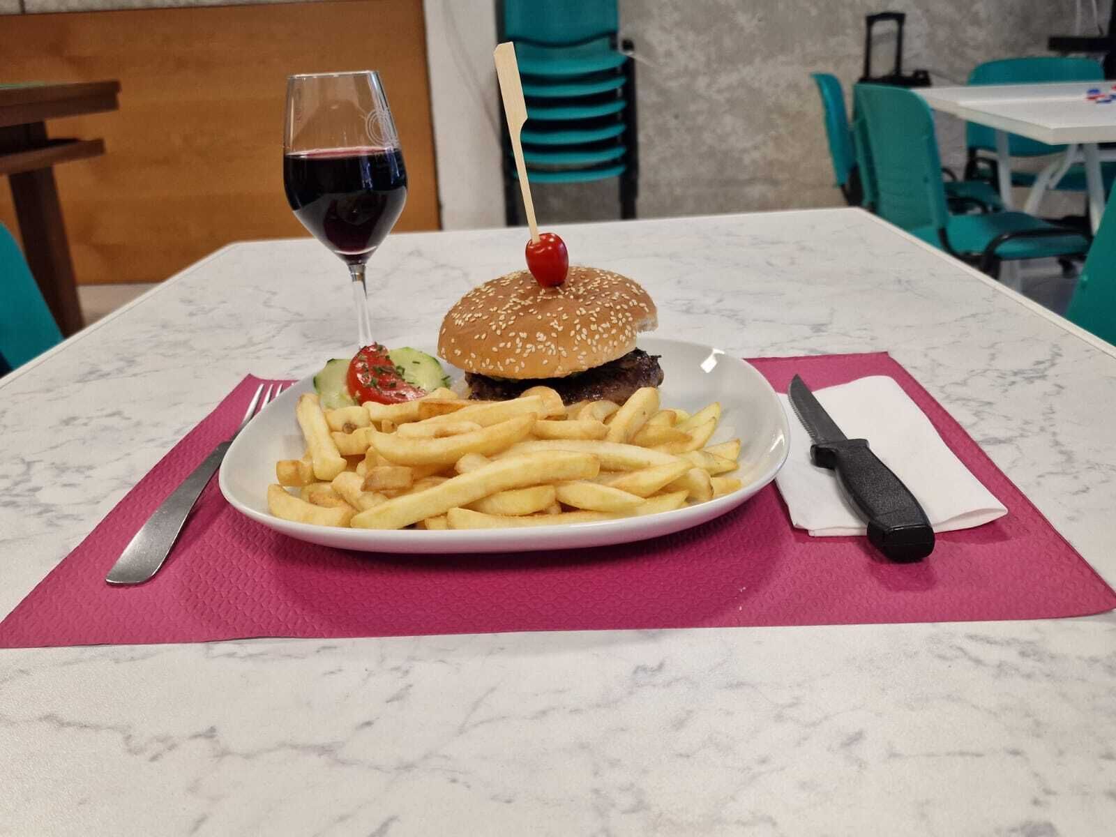 Una comida que incluye una hamburguesa con cereza encima, una copa de vino tinto y una porción de papas fritas sobre un plato blanco, colocada sobre una mesa con mantel individual rosa.