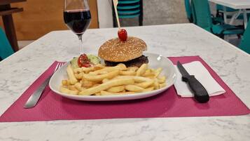 Una comida que incluye una hamburguesa con cereza encima, una copa de vino tinto y una porción de papas fritas sobre un plato blanco, colocada sobre una mesa con mantel individual rosa.