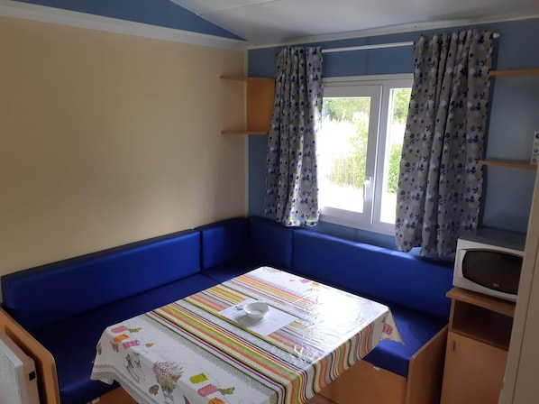 A small room with a bed, a window with curtains, a microwave, and a table with a striped tablecloth. - Camping les Lavandes (Castellane)