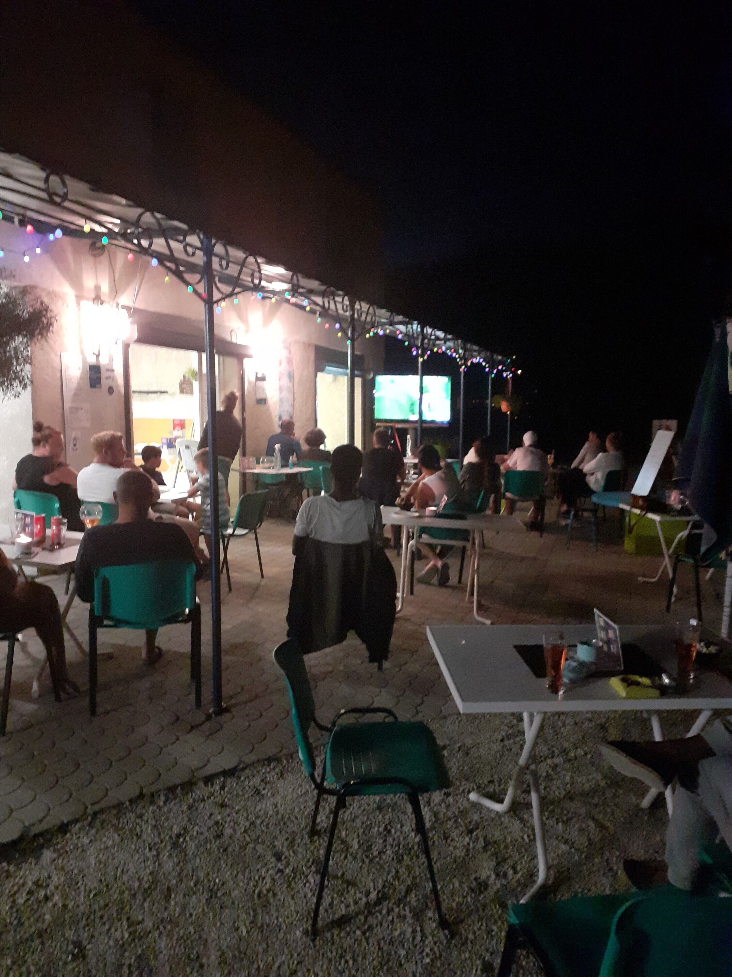 An outdoor seating area with tables and chairs, a television screen, and string lights.