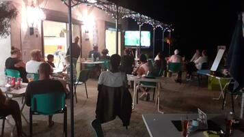 An outdoor seating area with tables and chairs, a television screen, and string lights.