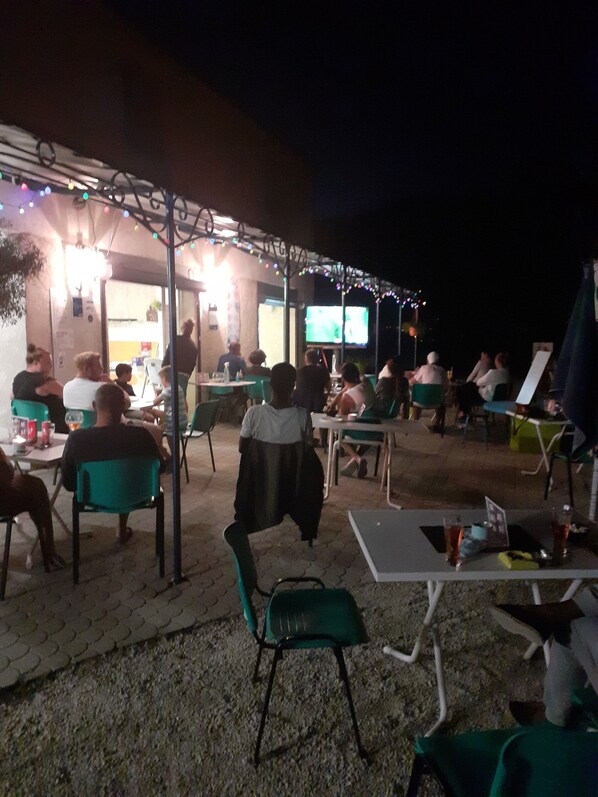 Un área de estar al aire libre con mesas y sillas, una pantalla de televisión y luces colgantes.