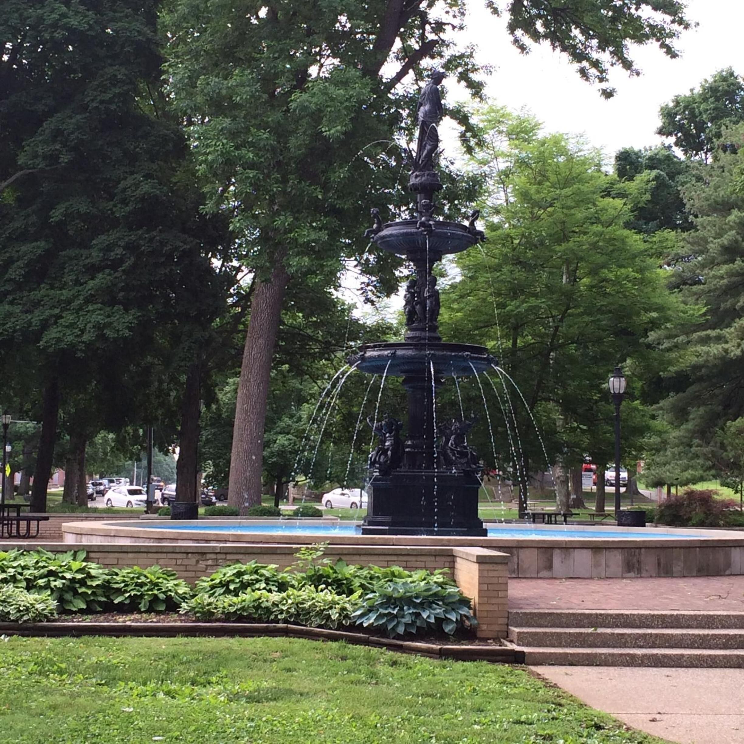 A park fountain with a statue base, surrounded by trees and a paved walkway.