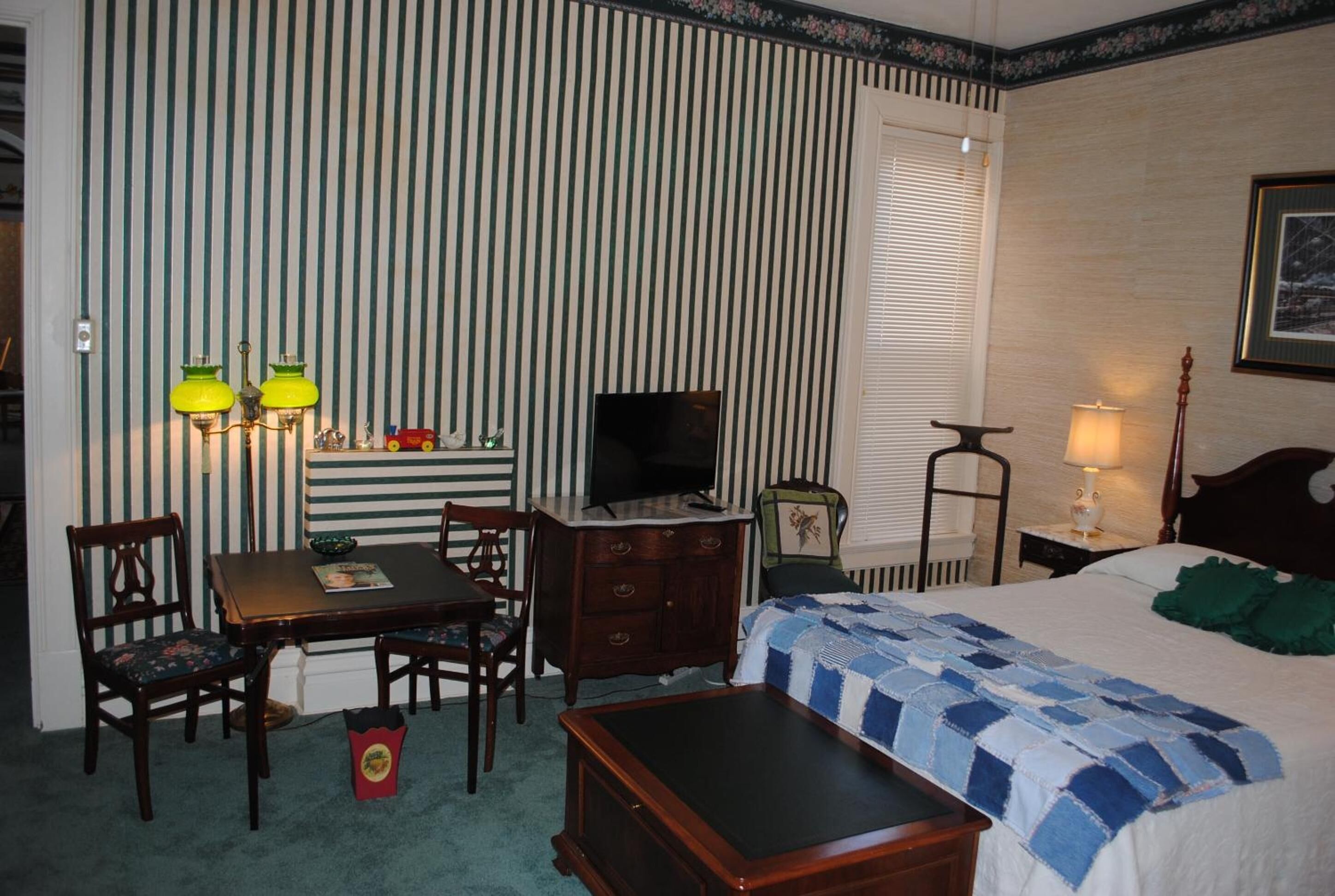 A hotel room with a striped wallpaper, a television, a desk with a lamp, a bed with a quilt, and a small table with chairs.