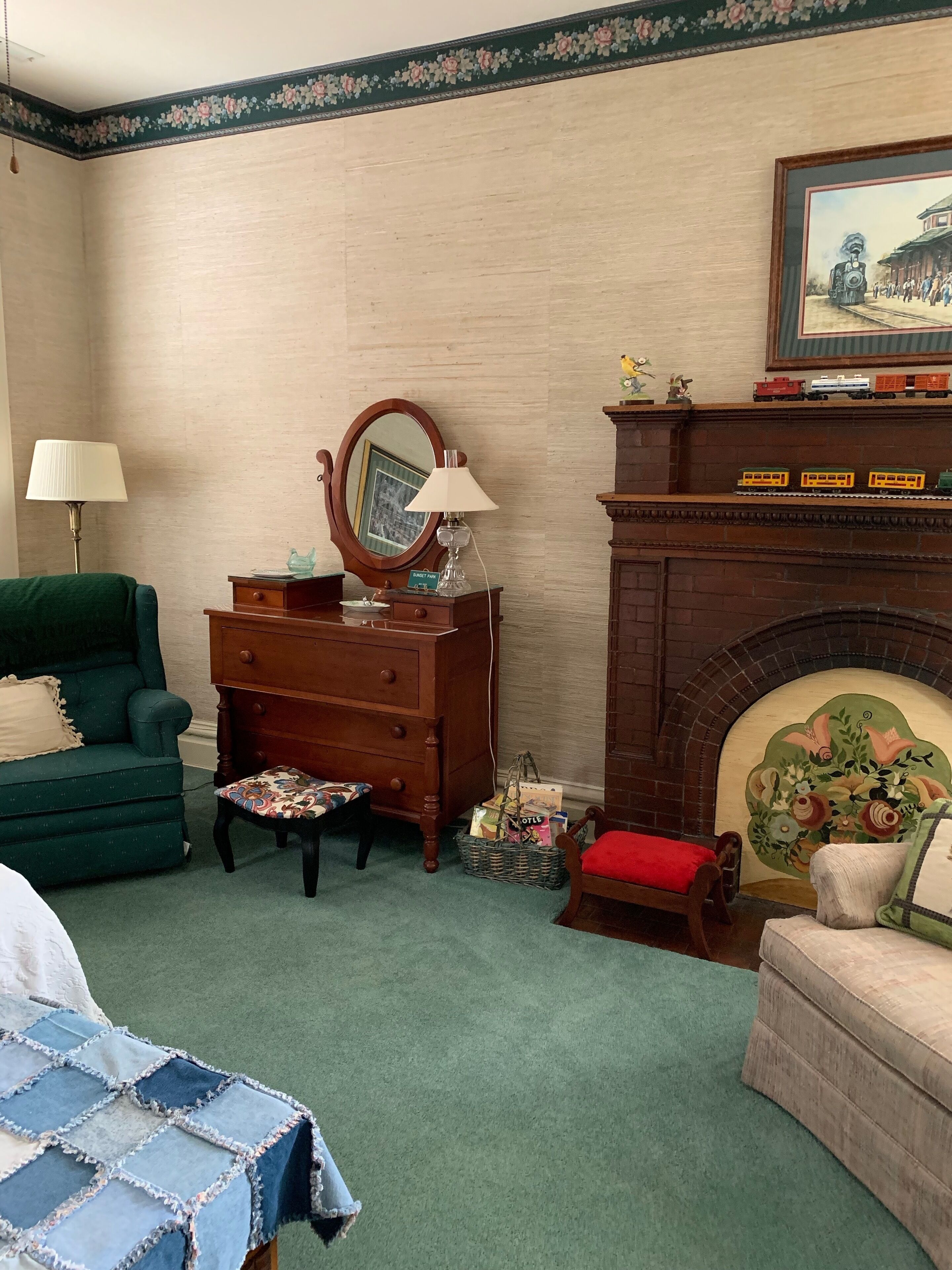 A room with a green armchair, a wooden dresser with a mirror, a small table with a lamp, a fireplace with a floral painting, and a bed with a blue quilt.