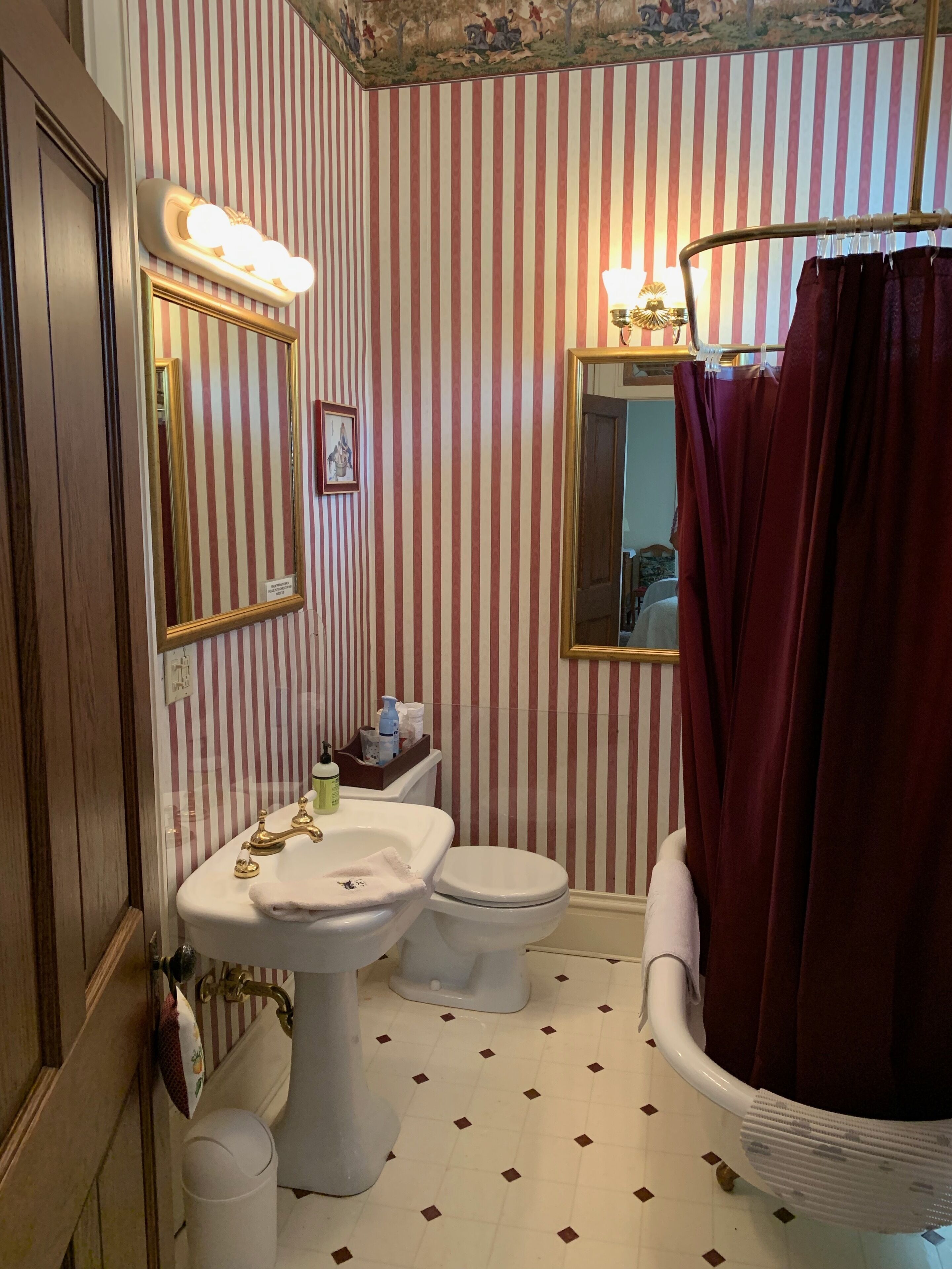 A bathroom with striped wallpaper, a white sink, a toilet, and a vintage-style bathtub with a red shower curtain.