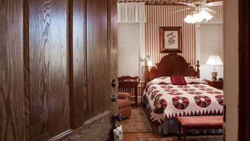 A bedroom with a wooden wardrobe, a bed with a floral quilt, a nightstand with a lamp, and a ceiling fan.