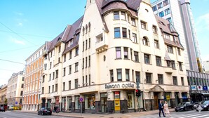 Un edificio de varios pisos con una entrada en esquina, una tienda visible llamada 'Fellmann Optika' y una calle con coches y peatones.