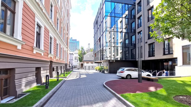 Una calle urbana moderna con un paseo adoquinado, edificios con grandes ventanales y un coche blanco estacionado al costado.