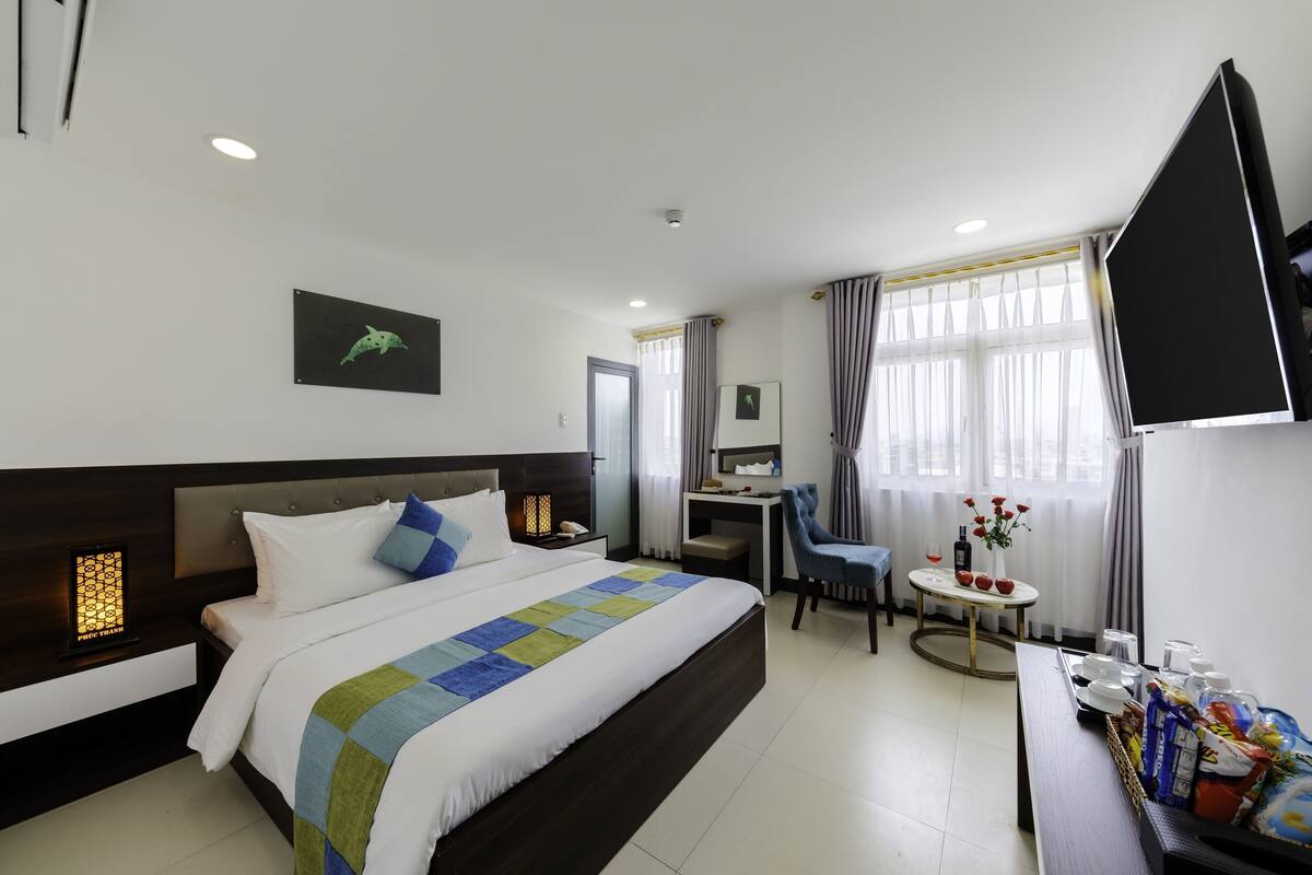 A hotel room with a large bed, a desk, a chair, a TV, and a small table with a vase of flowers.