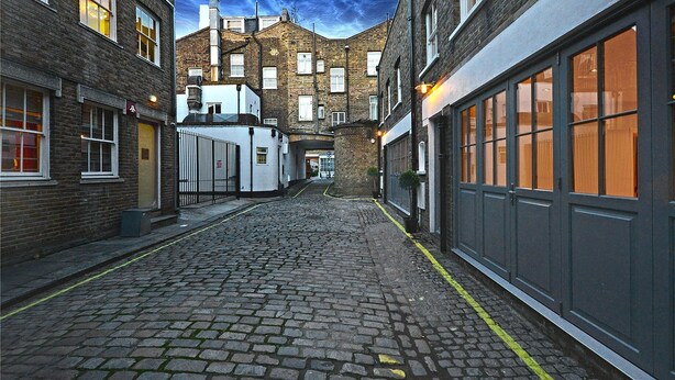 A cobblestone alleyway between buildings with brick facades, large windows, and a clear sky. - Rainsford Mews by Lime Street (London)