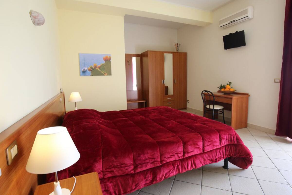 A hotel room with a bed, a desk with a chair, a wardrobe, a TV, and a painting on the wall.