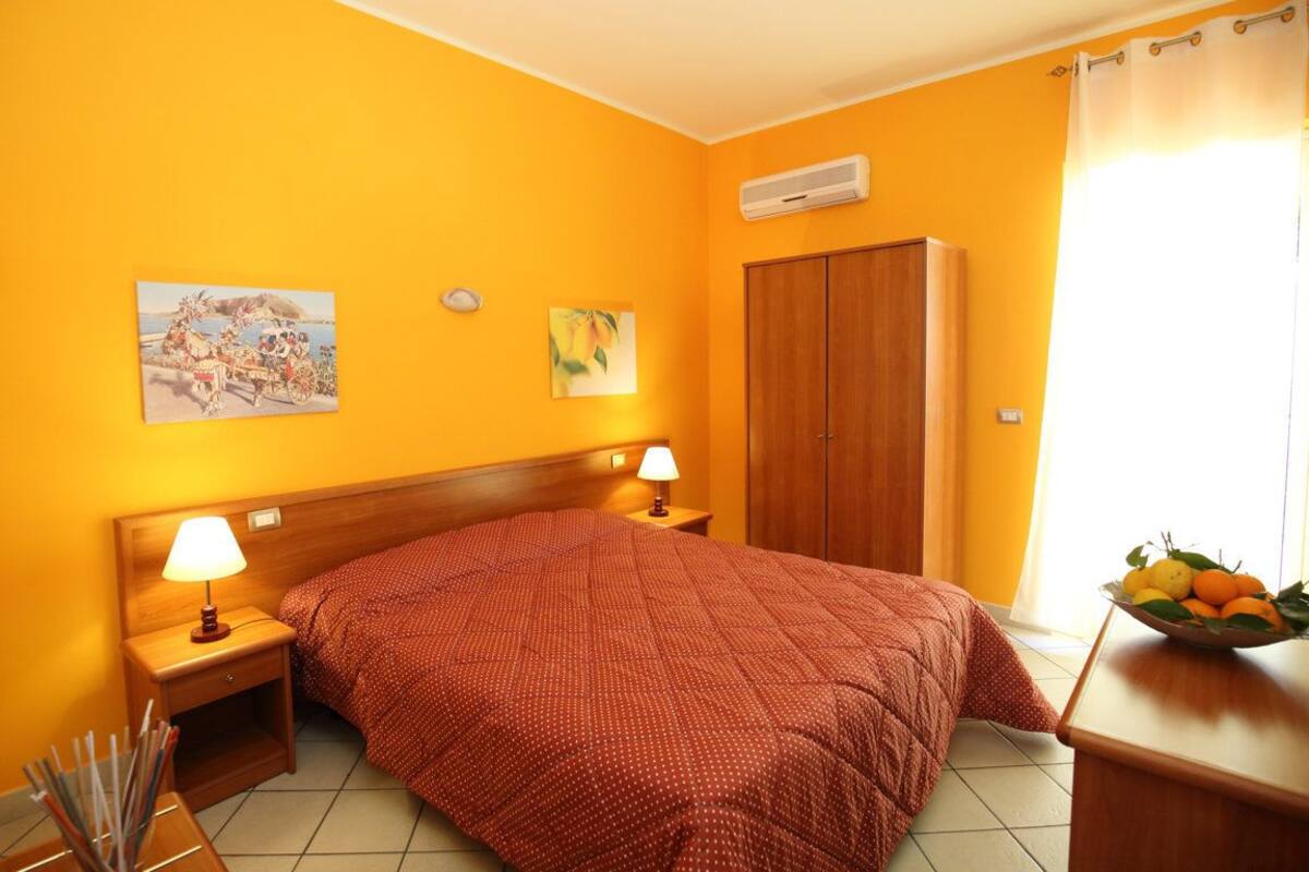 A hotel room with a large bed, wooden headboard, two bedside tables with lamps, a wardrobe, and artwork on the walls.