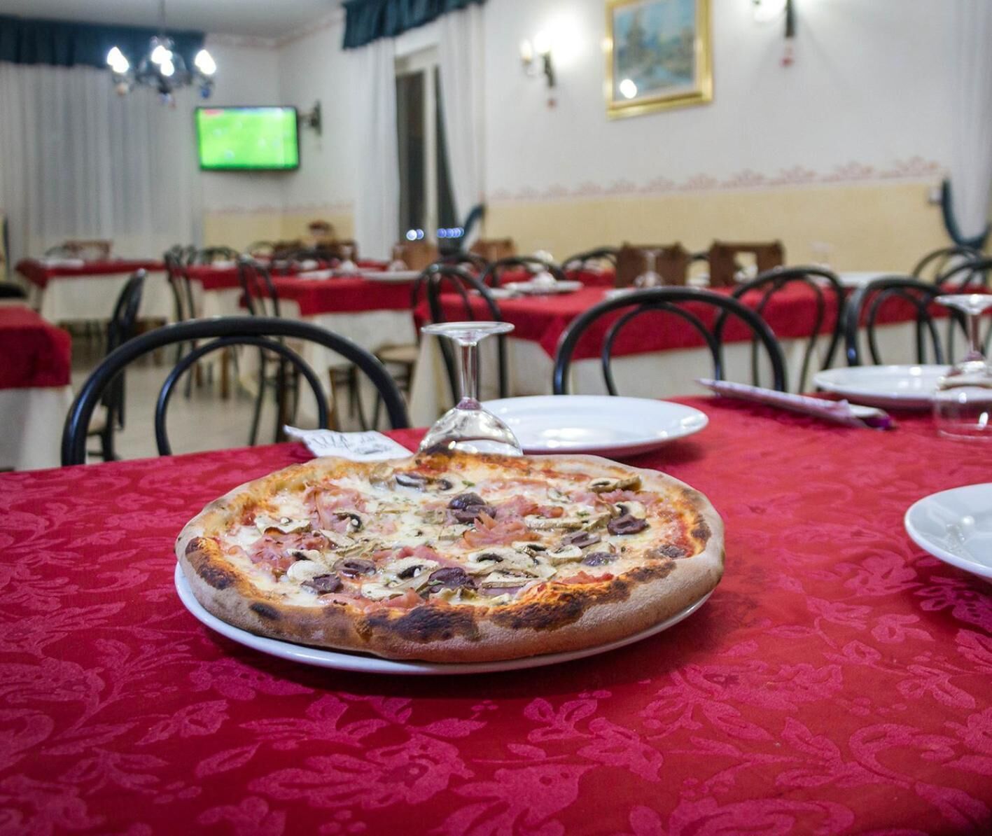 Une pizza sur une table recouverte d’une nappe rouge, dans un coin repas avec des chaises et des tables.