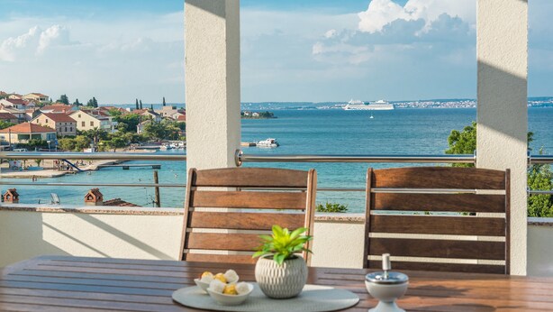 Un balcón con sillas de madera, una mesa con una planta y un cuenco de fruta, con vista a un pueblo costero y al mar.