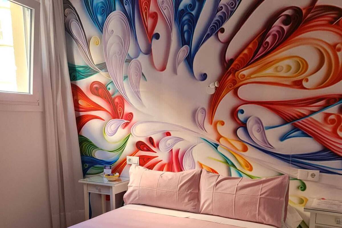 Un dormitorio con un mural abstracto y colorido en la pared, una cama con ropa de cama rosa y una mesita de noche pequeña con una lámpara.