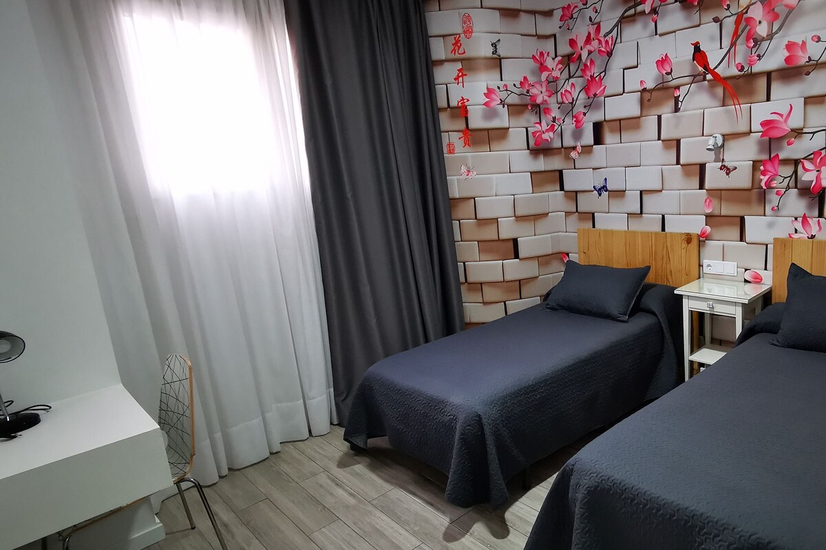 Habitación de hotel con dos camas individuales, una mesita de noche pequeña, un escritorio con lámpara y una pared decorada con motivos florales.