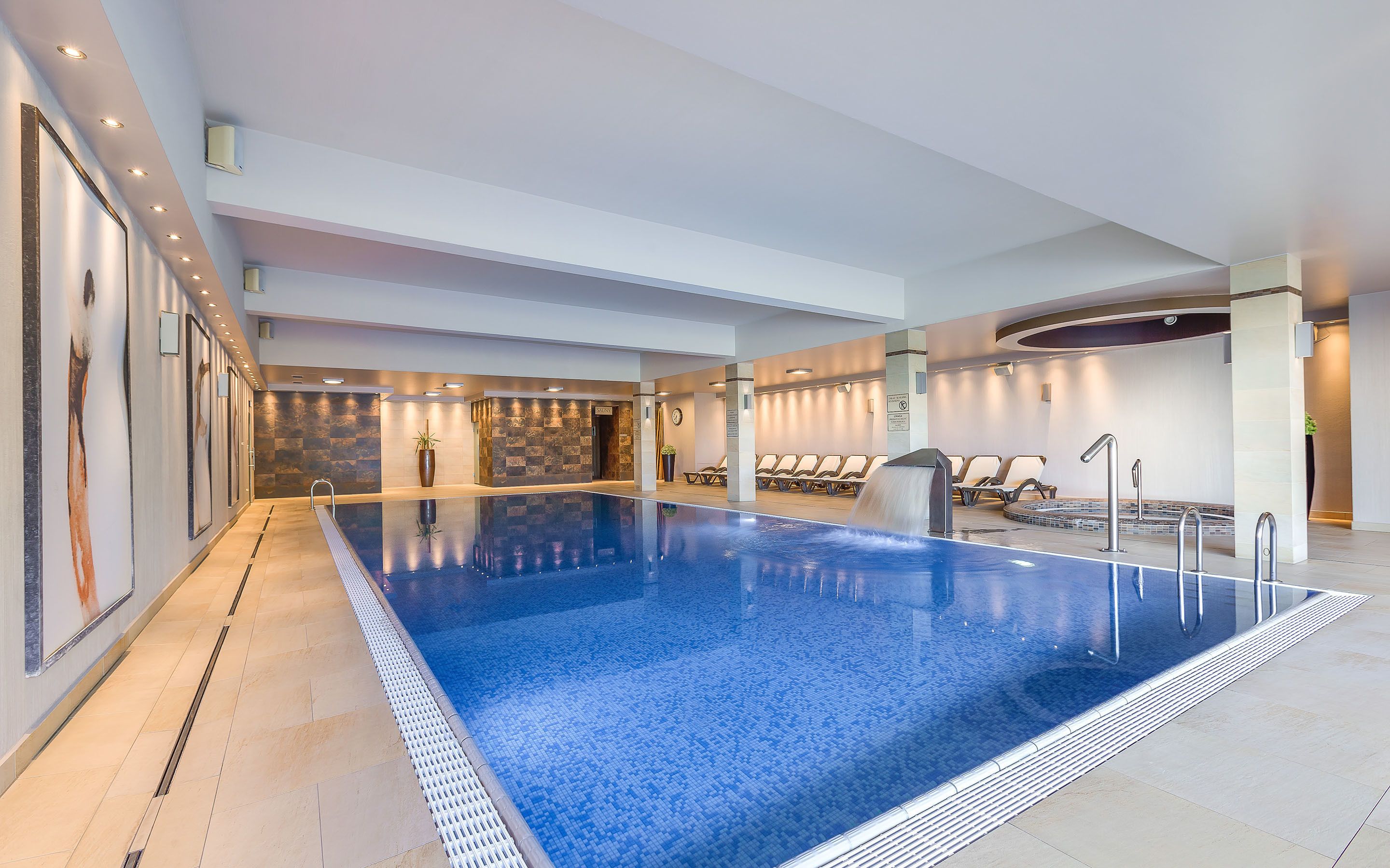 Una amplia piscina interior con suelo de baldosas, una zona de estar con tumbonas y una recepción.
