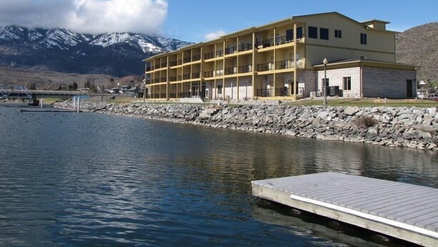 Un hotel de varios pisos con balcones, situado junto a un lago y montañas al fondo.